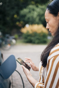 Side view of mature woman using smart phone while charging car in back yard