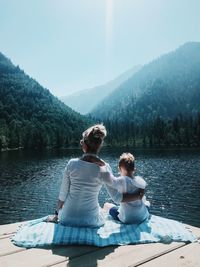 Rear view of mother and son with arm around sitting by lake against mountain