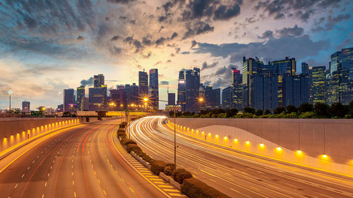 Light trails on road amidst buildings against sky during sunset