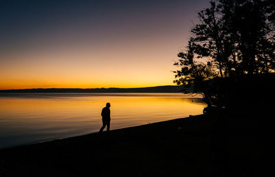 Silhouette man walking on lakeshore against sky during sunset