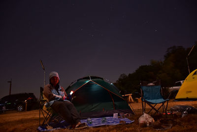 Rear view of man sitting at illuminated tent against sky at night