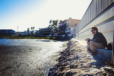 Man sitting on retaining wall in city against clear sky