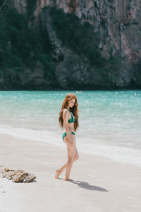 Full body side view of fit female in swimwear standing on sandy coast near azure sea against mountain in thailand
