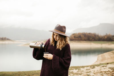 Man holding hat standing by lake against sky