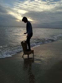 Side view of boy standing on chair at beach against sky during sunset