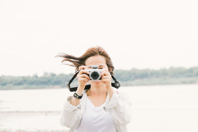 Woman photographing using camera against lake and clear sky