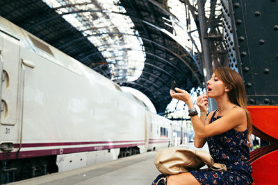 Beautiful woman applying make-up while sitting on platform at railroad station
