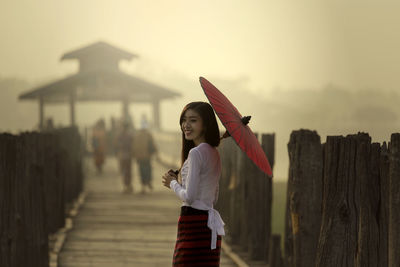 Young woman with umbrella standing on pier against sky