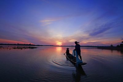 Silhouette men on rowboat in lake against dramatic sky during sunset