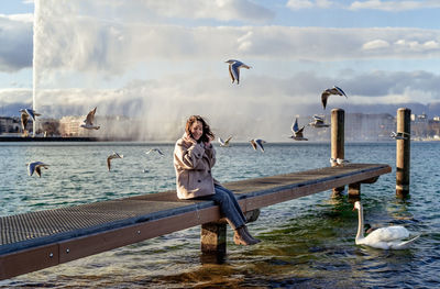 Happy smiling woman sitting on pier on lake with seagulls and swan enjoying fresh breeze jet d'eau