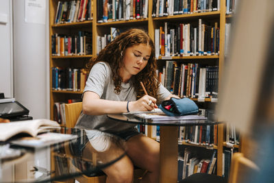 Focused curly haired teenage girl writing while sitting near table in library at junior high school