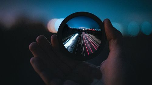 Close-up of hand holding lens over light trails on road at night