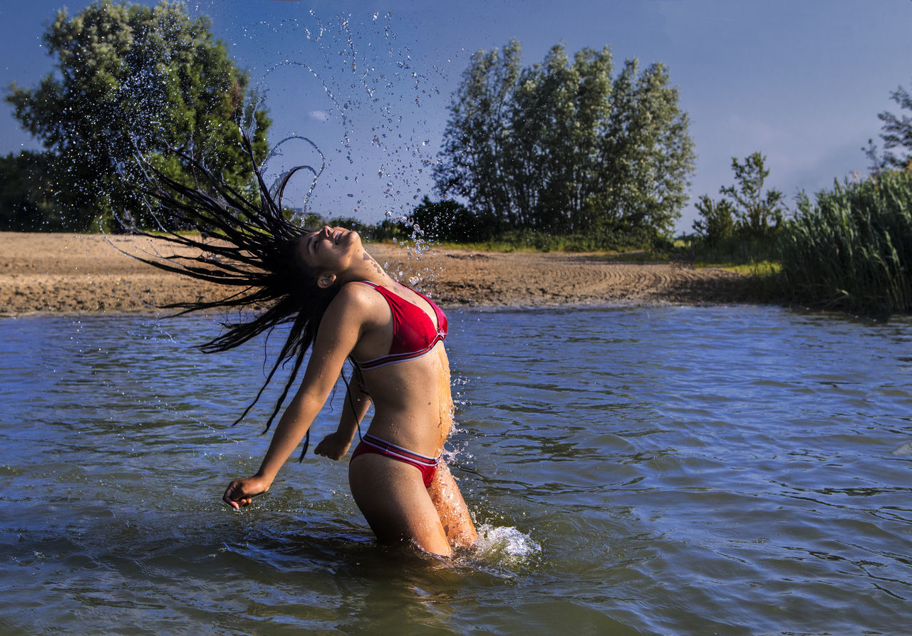 Girl in bikini splashing water in lake while tossing hair against sky