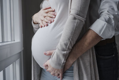 Pregnant woman with husband's hands on stomach standing by window at home