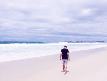 Rear view of mid adult man walking on beach against cloudy sky