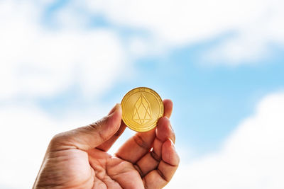 Close-up of cropped hand holding coin against cloudy sky