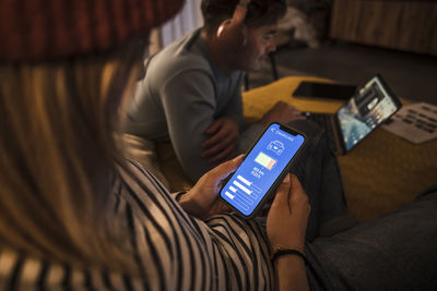 Young woman operating electric car charging app on smart phone and man using laptop in background