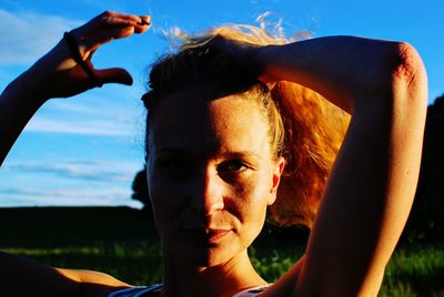 Portrait of young woman tying hair against sky during sunset