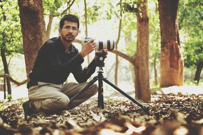 Portrait of young photographer with tripod camera on field by trees at park