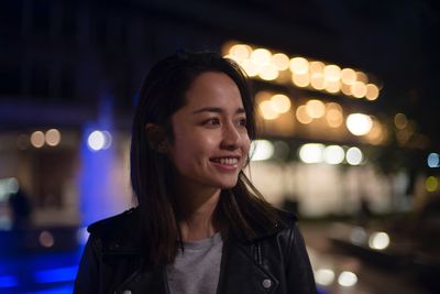 Smiling young woman looking away against illuminated buildings in city