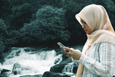 Side view of woman using mobile phone by waterfall in forest