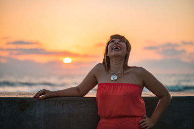 Woman laughing standing by retaining wall against cloudy sky during sunset