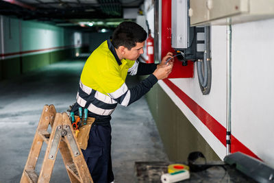 Side view of young professional male technician with electric tools repairing and checking equipment while working in building