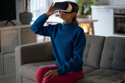 Carried away european woman in virtual reality headset looking up using vr and ar sits on sofa