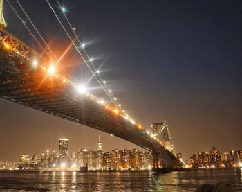 Illuminated bridge over river by buildings against sky at night
