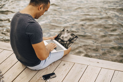 Mature man using laptop while sitting on jetty over lake
