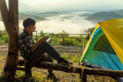 Full length of young man drinking coffee while using mobile phone by tent in forest