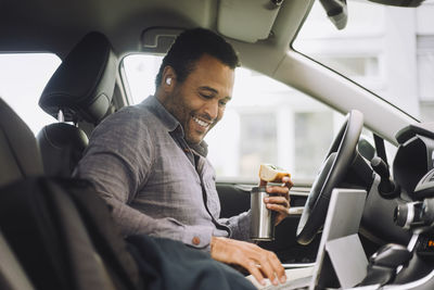 Smiling businessman with insulated drink container using laptop while sitting in car