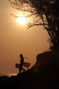 Silhouette fisherman carrying baskets while walking on rock formation at beach during sunset