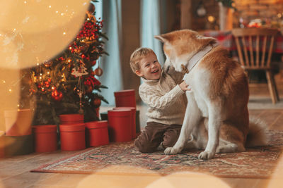 Candid authentic happy little boy in knitted beige sweater hugs dog with bow tie at home on xmas