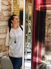 Young woman looking away while standing against window