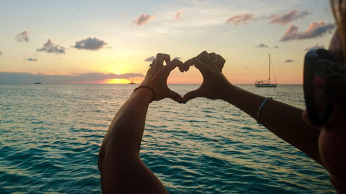 Cropped hands making heart shape by sea against sky during sunset