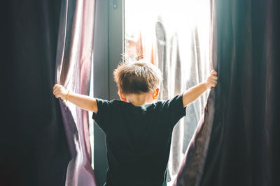 Rear view of boy with arms outstretched opening curtan while standing against window