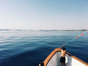 Rear view of shirtless man standing in boat on sea against clear sky