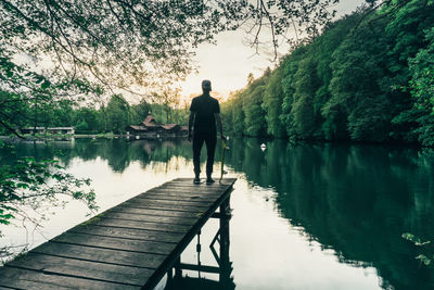 Rear view of boy with skateboard standing on pier over lake against trees