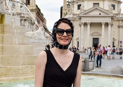 Portrait of smiling woman wearing sunglasses against fountain in town