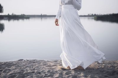 Low section of woman wearing white dress standing by lake