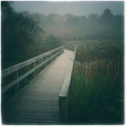 Wooden footbridge over grassy field against sky during foggy weather