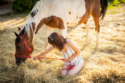 Woman sitting by horse grazing on grass