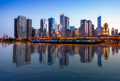 Illuminated buildings by lake in city against sky at dusk