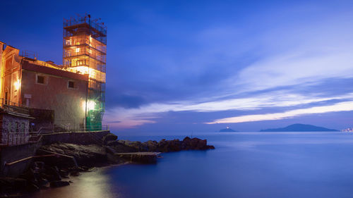 Illuminated buildings by sea against sky at sunset