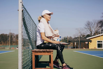 Side view of tired senior woman sitting on bench against sky at tennis court