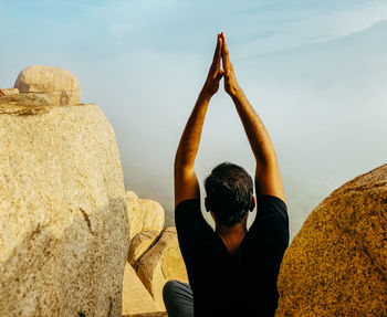 Rear view of man sitting on rock and doing yoga against sky