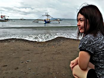 Side view of smiling young woman crouching on beach against cloudy sky