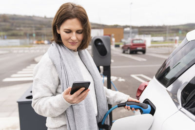 Woman using smart phone charging electric car at station