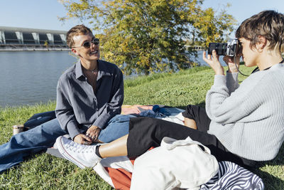 Young woman photographing smiling friend through camera while sitting at park